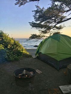 there is a tent set up on the beach by the fire pit and trees near the water