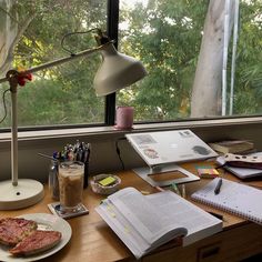 a desk with an open book, notebooks and a lamp on it next to a window