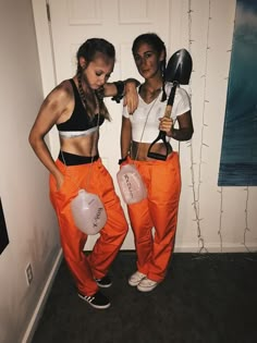 two young women dressed in orange pants and white shirts, one holding a pair of scissors