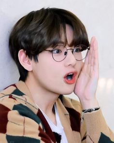 a young man wearing glasses making a hand gesture with his right hand while standing against a wall