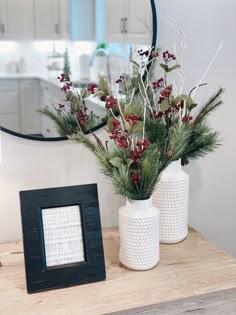 two white vases with red berries and greenery sit on a table in front of a mirror