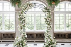 an arch decorated with white flowers and greenery in front of two windows at the end of a hall