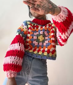 a woman wearing a colorful crochet sweater with her hands in the air while standing next to a white wall