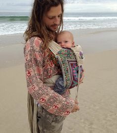 a man with long hair holding a baby in his arms while standing on the beach