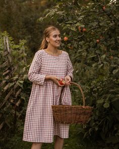 a woman standing in an apple orchard holding a wicker basket full of ripe apples