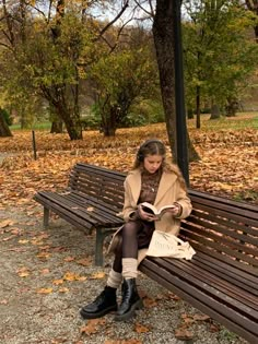 a woman sitting on a bench reading a book in a park with leaves all over the ground