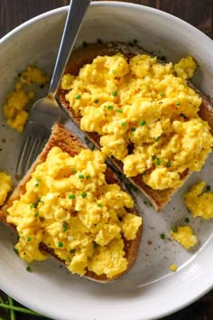 two pieces of toast with scrambled eggs on them and a fork in the bowl next to it