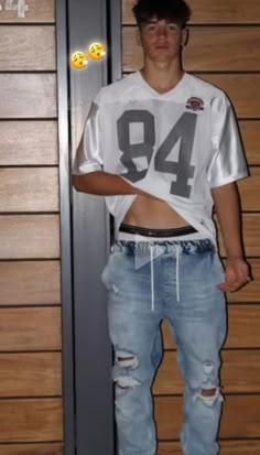 a young man standing in front of a wall wearing ripped jeans and a football jersey