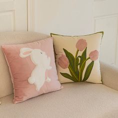 two pillows on a couch with flowers and a bunny sitting in the center one is pink