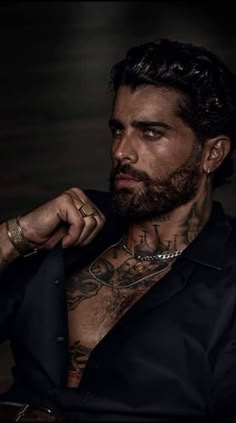 a man with tattoos on his chest wearing a black shirt and gold bracelets sitting in a chair