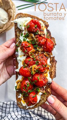 someone holding up a toast with tomatoes and goat cheese on it, topped with fresh herbs