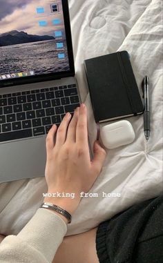 a person using a laptop computer on top of a bed with a book and pen