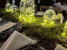 the table is decorated with glass jars and fairy lights on moss covered plates, napkins, and silverware