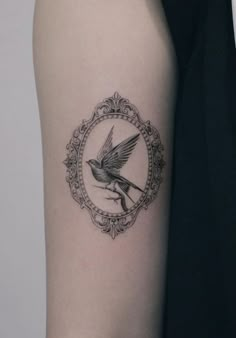 a small bird tattoo on the left upper arm and lower arm, with an ornate frame around it