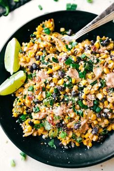 a black plate topped with corn salad next to lime wedges and a fork on the side