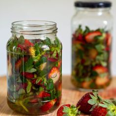 two jars filled with strawberries and greens on top of a wooden table next to each other