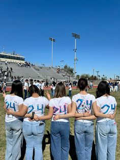 four girls in white shirts and blue jeans are standing on the field with their arms around each other