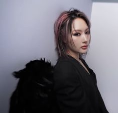 a woman with pink hair and black jacket leaning against a white wall, looking at the camera