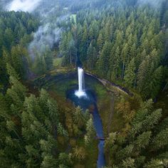 an aerial view of a waterfall in the middle of a forest with fog coming from it