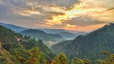 the sun is setting over some mountains and trees in the foreground, with mist rising from the valley below