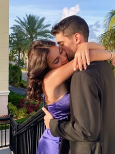 a man and woman embracing each other in front of a fence with palm trees behind them