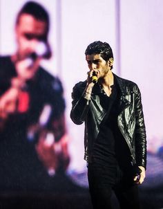 a man in black leather jacket standing on stage holding a microphone to his mouth and singing