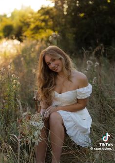 a woman in white dress sitting on grass and smiling at the camera while holding flowers