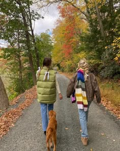 two people walking down a road with a dog on the other side and trees in the background