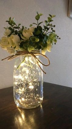 a mason jar filled with white flowers and string lights on a wooden table next to a wall