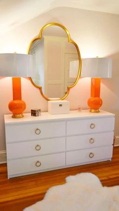 a white dresser with two orange lamps and a gold mirror on it's top