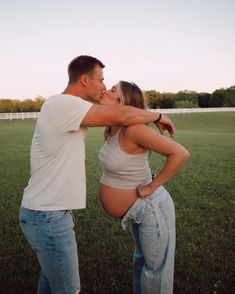 a man and woman are kissing in the middle of a grassy field with a white fence behind them