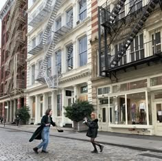 two people are walking down the street in front of tall buildings with fire escapes on them