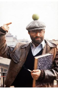 a man with a book pointing at an apple on top of his hat and jacket