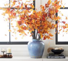 a blue vase filled with yellow and red leaves on top of a white table next to a window