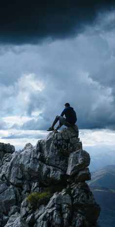 a man sitting on top of a large rock next to a mountain under a cloudy sky