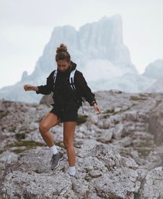 a woman in black jacket and shorts standing on rocks with mountains in the back ground