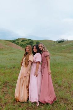 three women in long dresses posing for the camera on a grassy field with hills in the background