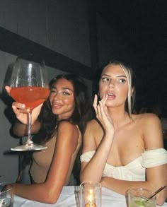 two beautiful women sitting at a table with wine glasses in front of them and one holding a drink