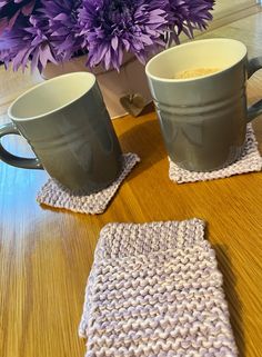 two coffee mugs sitting on top of a wooden table next to a purple flower