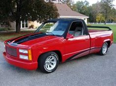 a red pick up truck parked in a parking lot next to a house and trees