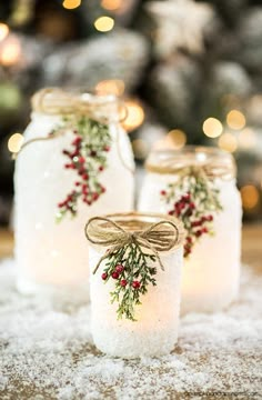 three mason jars decorated with holly and red berries are sitting on snow covered ground in front of a christmas tree