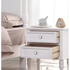 a white nightstand table with two drawers and a clock on the top, in front of a bed