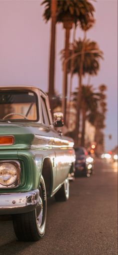 an old green car is driving down the street with palm trees in the back ground