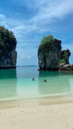 two people are swimming in the clear blue water near large rock formations on an island