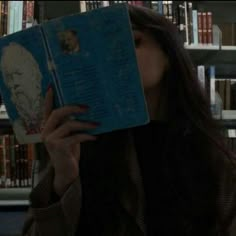 a woman holding up a book in front of her face with bookshelves behind her