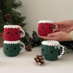 crocheted coffee mug cozyies are being held by two hands with pine cones on the table