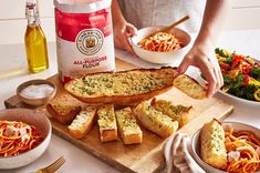 a person is preparing food on a cutting board with bread, pasta and salad in bowls