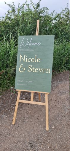 a wooden sign sitting on top of a gravel road next to tall green grass and bushes