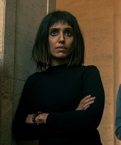 a woman standing next to a wall with her arms crossed and looking at the camera