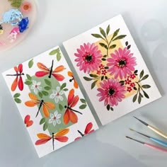 two cards with flowers on them next to paintbrushes and watercolors in the background